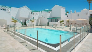 En swimmingpool med et glashegn, omgivet af hvide bygninger med altaner og palmer.