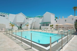 A swimming pool with a glass enclosure, surrounded by white buildings with balconies and palm trees.