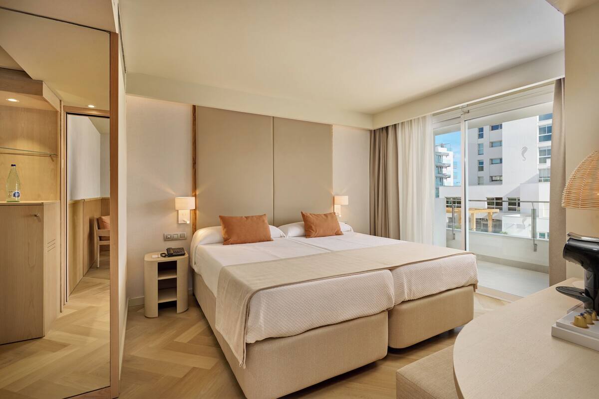 Ein modernes Hotelzimmer mit einem großen Bett, Nachttischlampen, einem kleinen Tisch und Blick auf die Stadt vom Balkon aus.