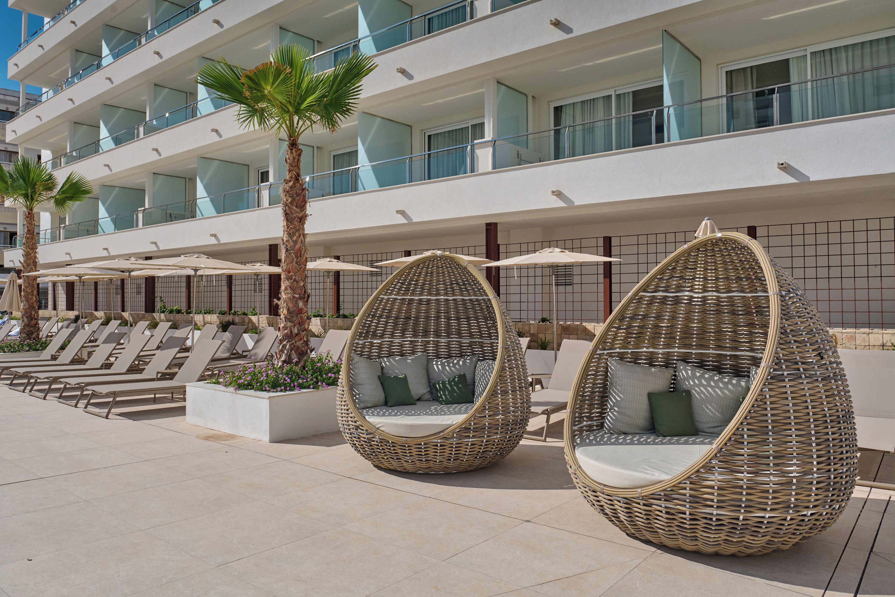 Outdoor wicker chairs with cushions, a poolside area, and a modern building with balconies.