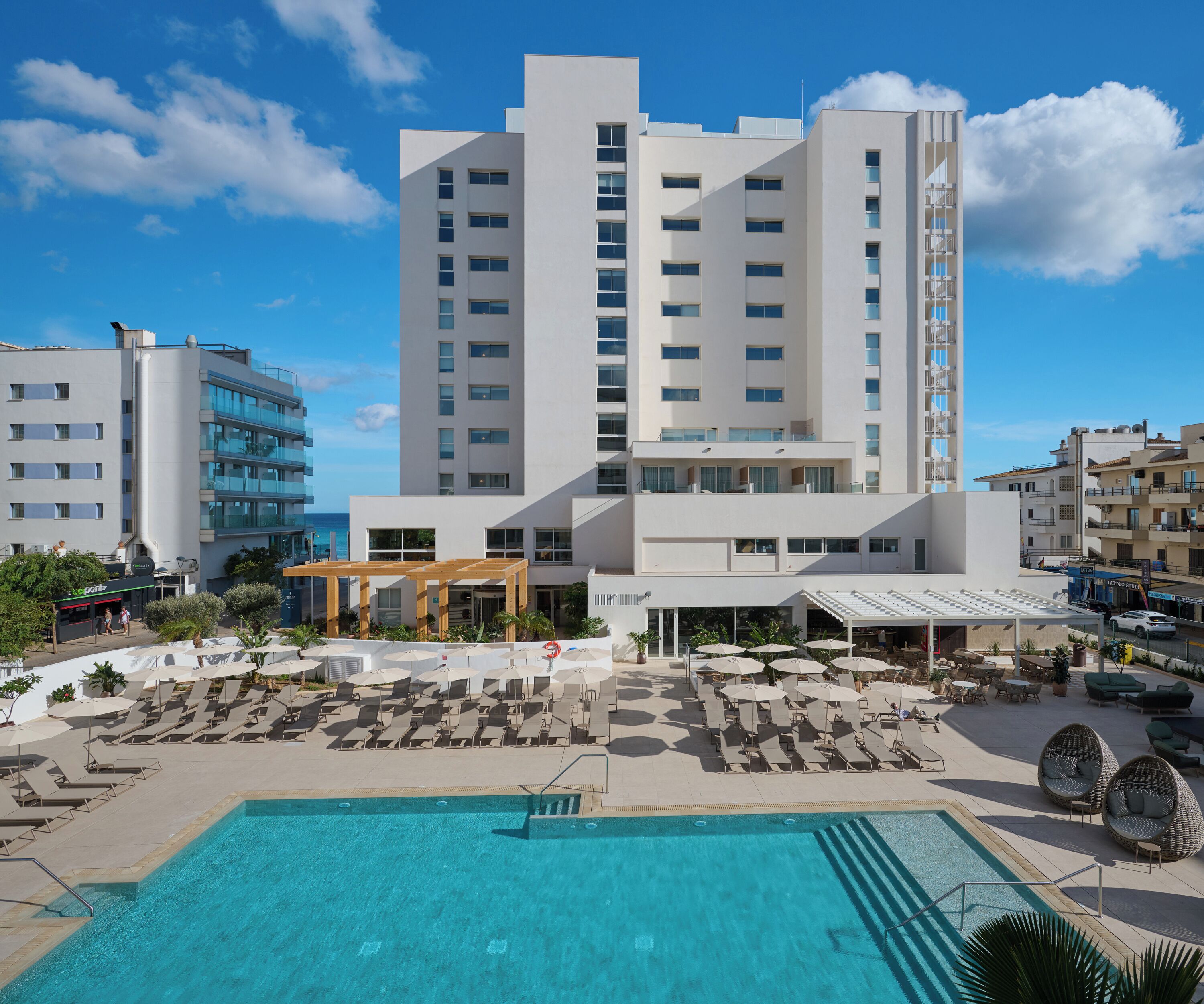 A modern hotel with a pool area, multiple floors, and a clear view of the sea.