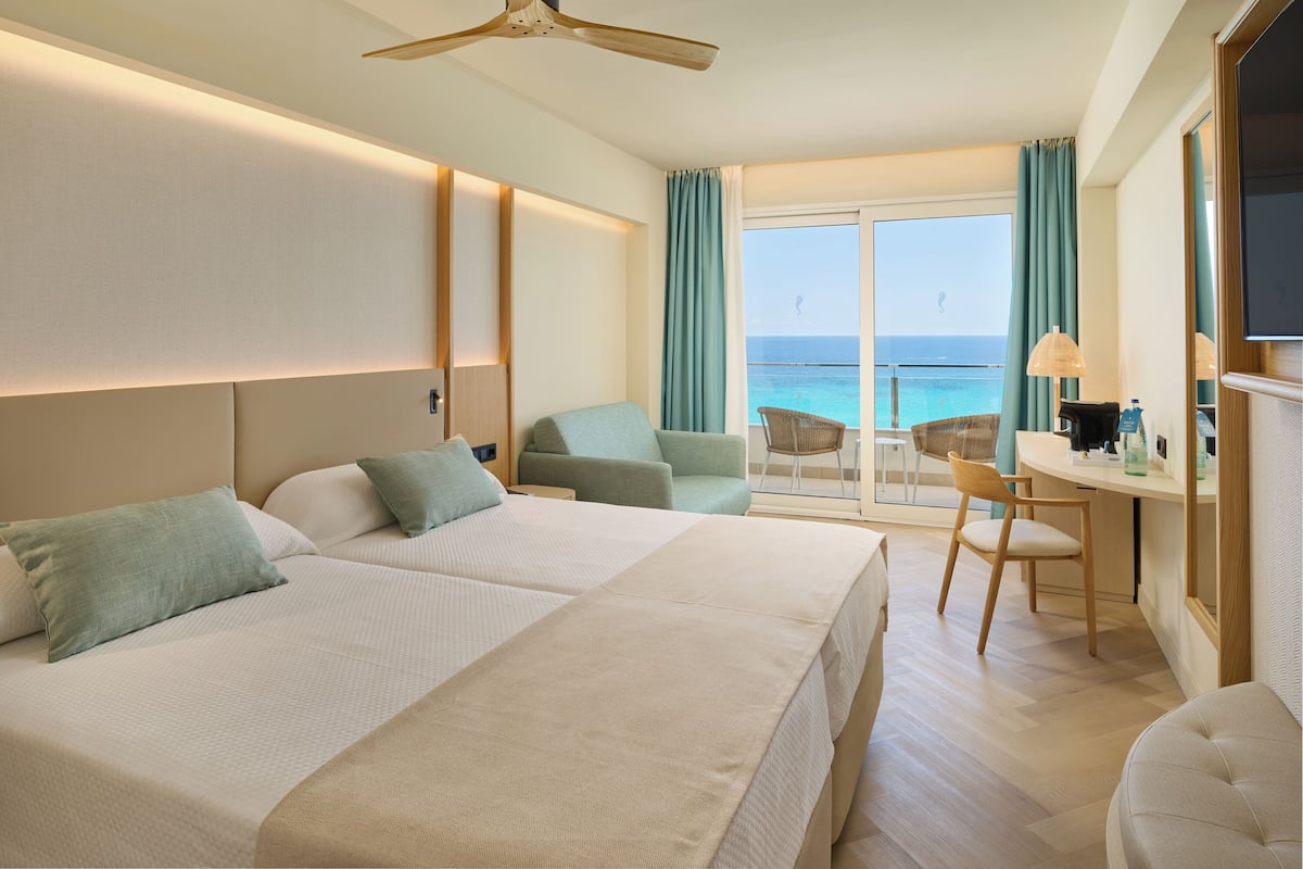 Ein Hotelzimmer mit zwei Betten, einem Schreibtisch, einem Sessel, einem Fernseher und einem Balkon mit Meerblick.