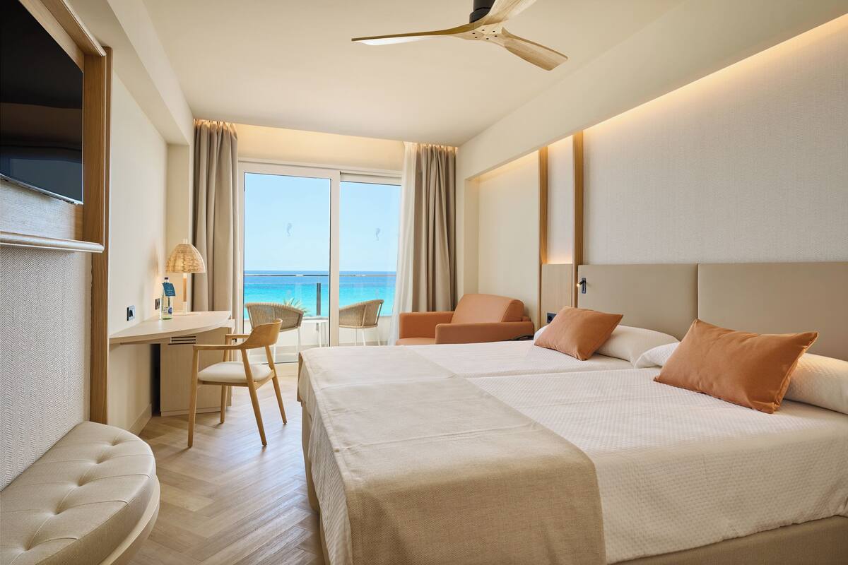 Ein Hotelzimmer mit einem großen Bett, einem Schreibtisch, einem Sessel und Blick auf das Meer.