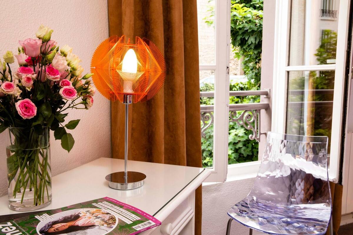 A room with a white table, a vase of pink and white roses, a clear glass chair, and a lit orange lamp.
