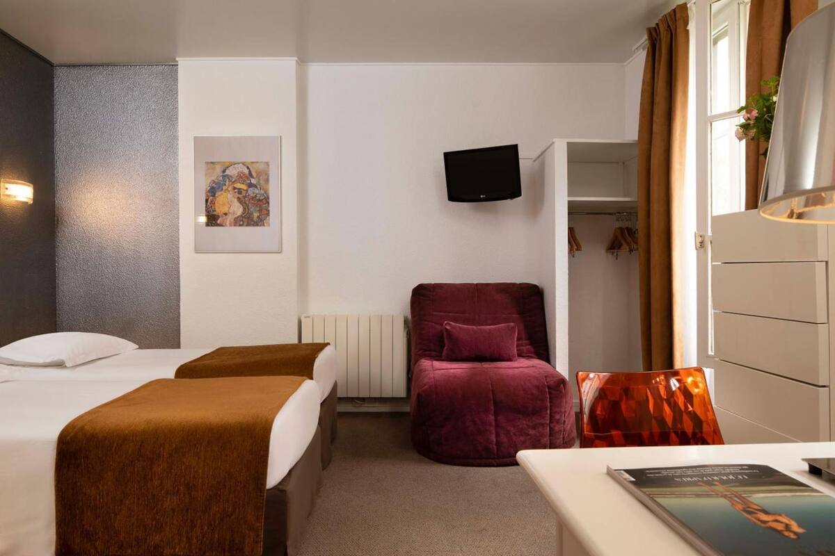 A hotel room with two beds, a red armchair, a TV, a radiator, and a wardrobe.