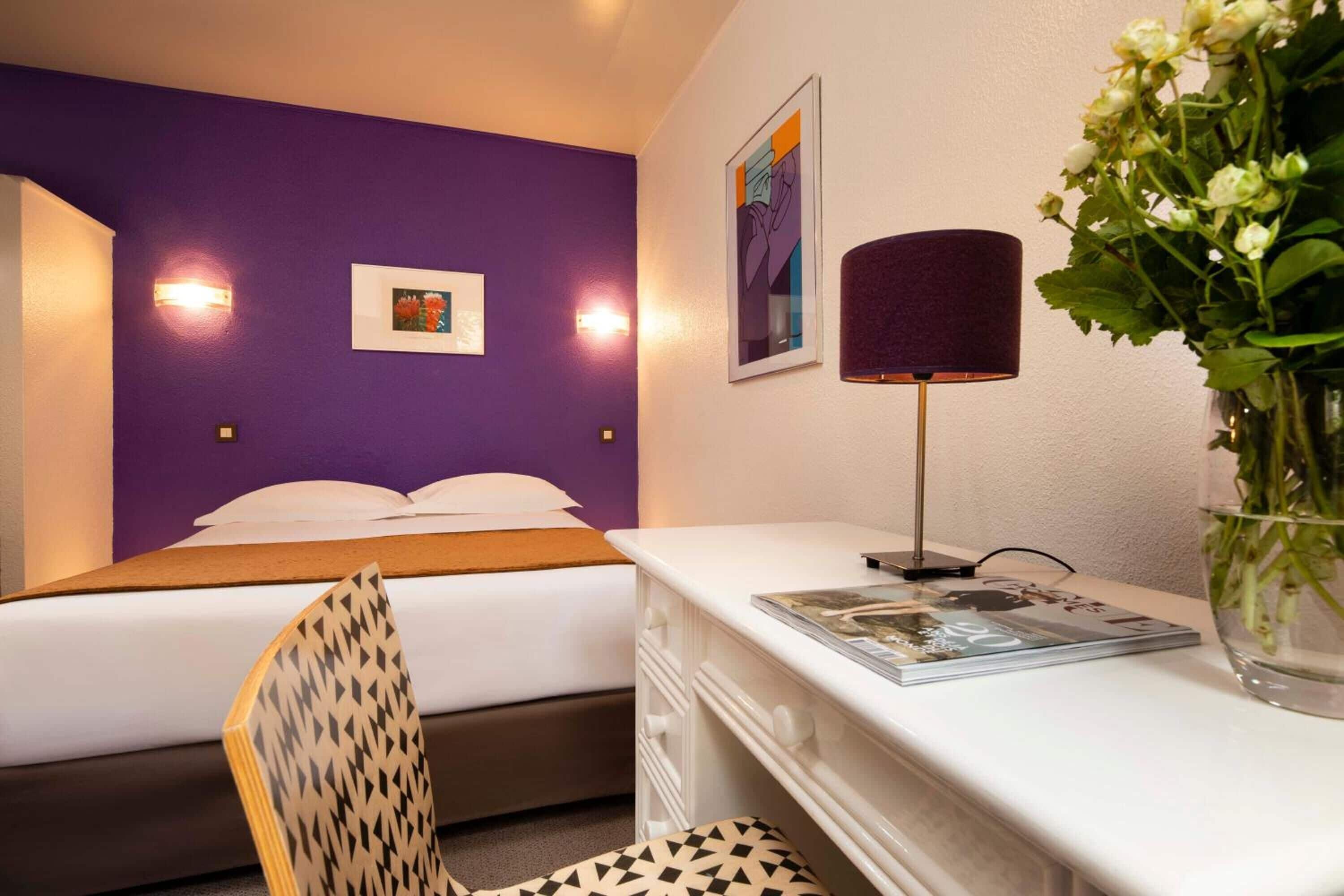 Habitació d'hotel amb una paret morada, un llit amb roba de llit blanca, una taula amb una làmpada i una cadira.