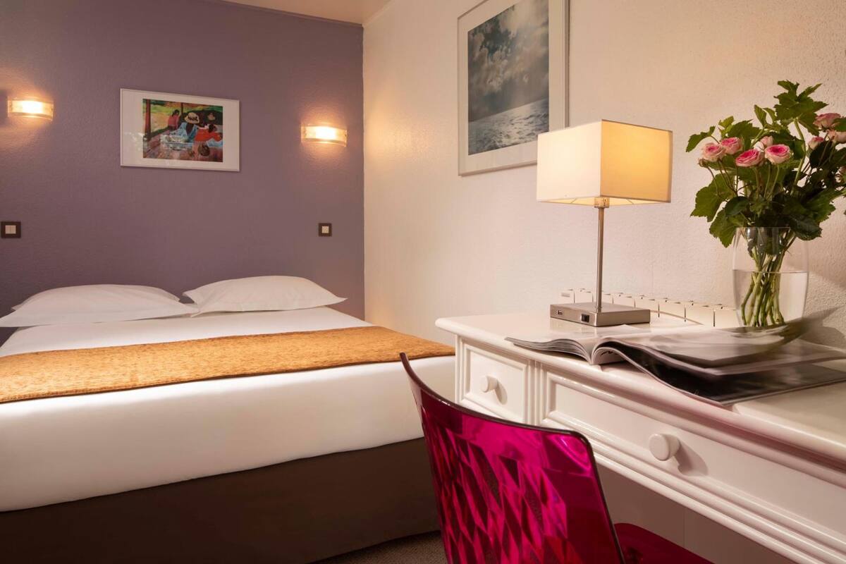 A hotel room with a bed, a desk with a lamp, a vase with flowers, and two wall-mounted lights.
