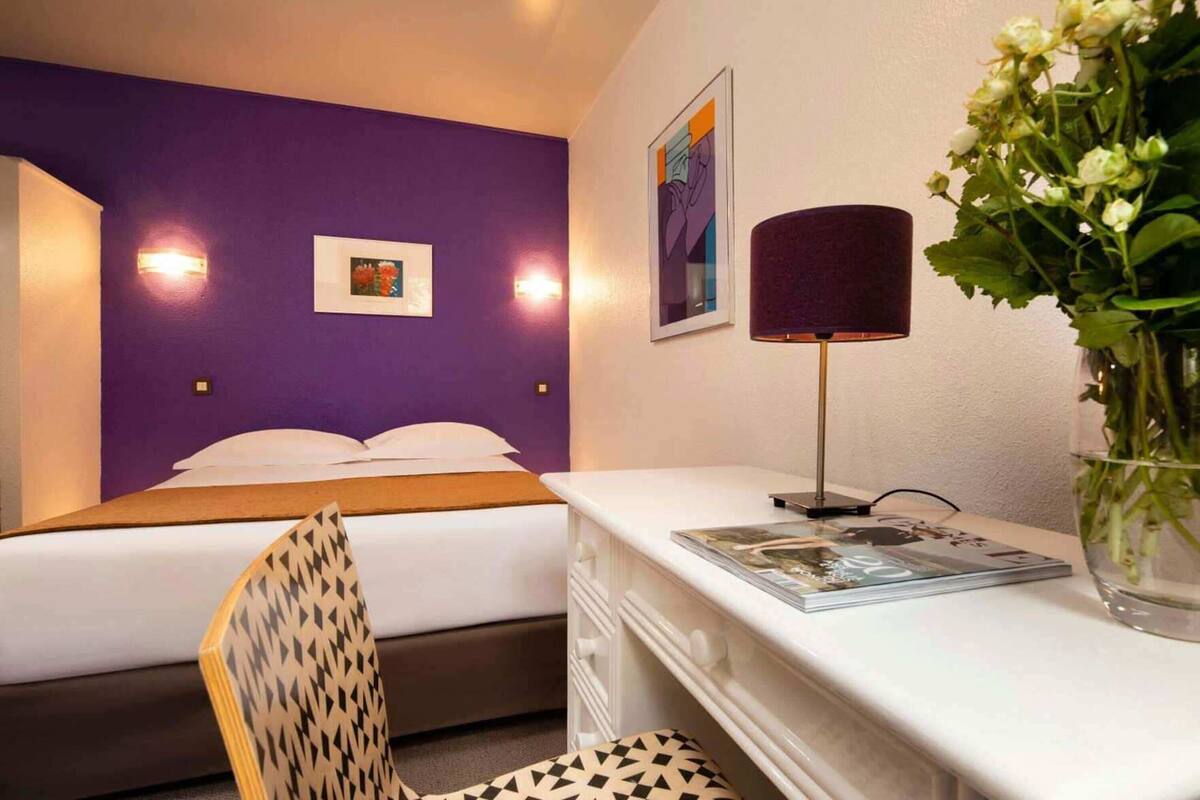 A hotel room with a bed, a desk with a lamp, a chair, and a vase with flowers.