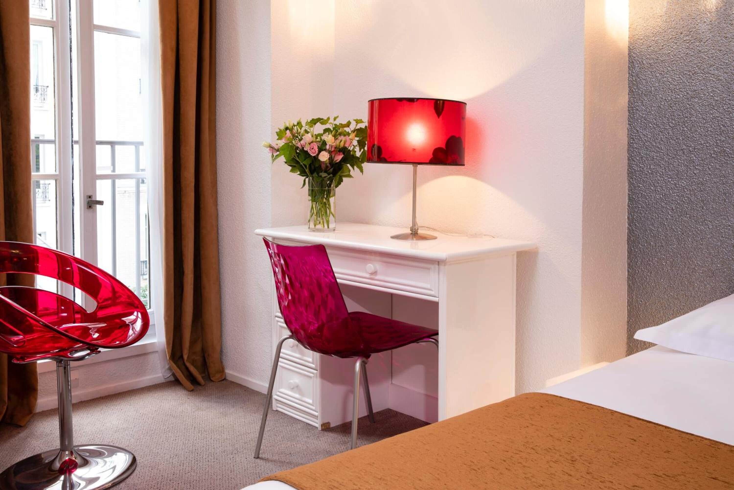 Una camera d'albergo con una scrivania bianca, una sedia rossa, una lampada rossa e un letto.