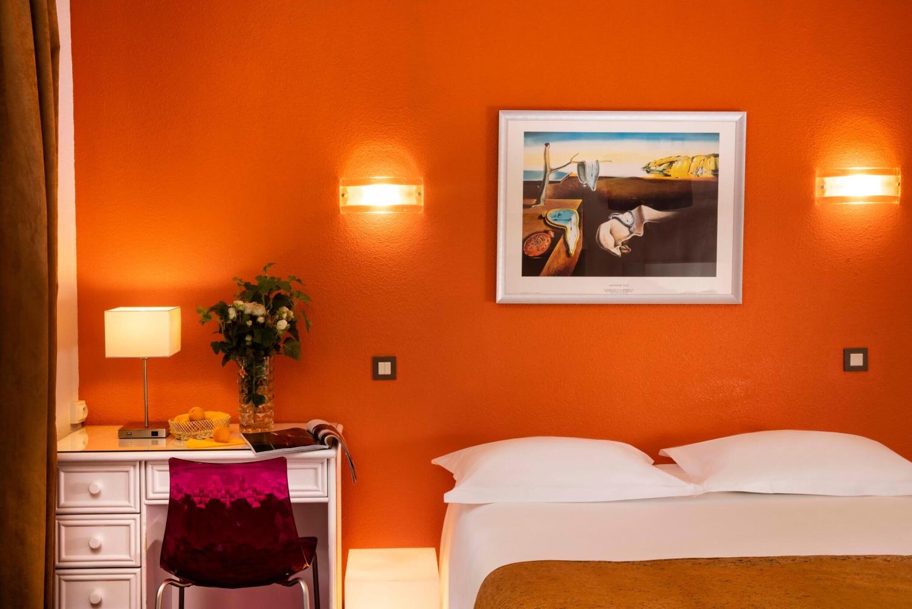 Habitación de hotel con una cama, un escritorio con lámpara, una silla, un florero con flores y un cuadro en la pared.