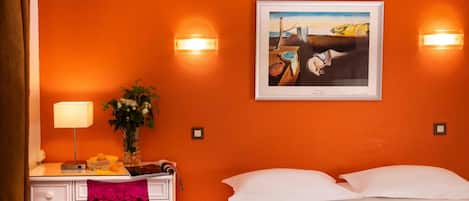 Habitación de hotel con una cama, un escritorio con lámpara, una silla, un florero con flores y un cuadro en la pared.