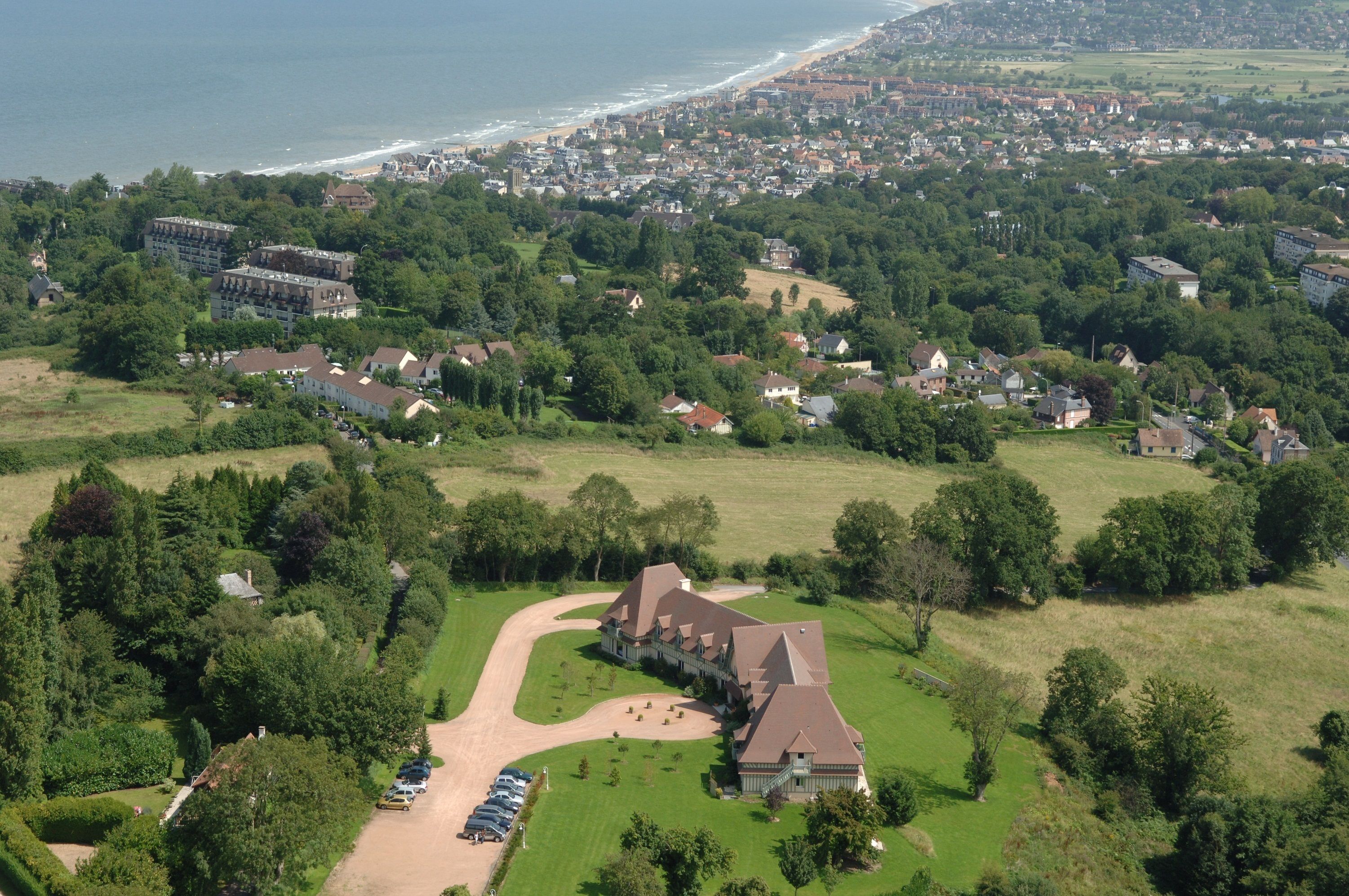 Ein Luftbild eines Wohngebiets mit einem großen Haus, umgeben von viel Grün, einem Strand im Hintergrund und einer kleinen Parkfläche.