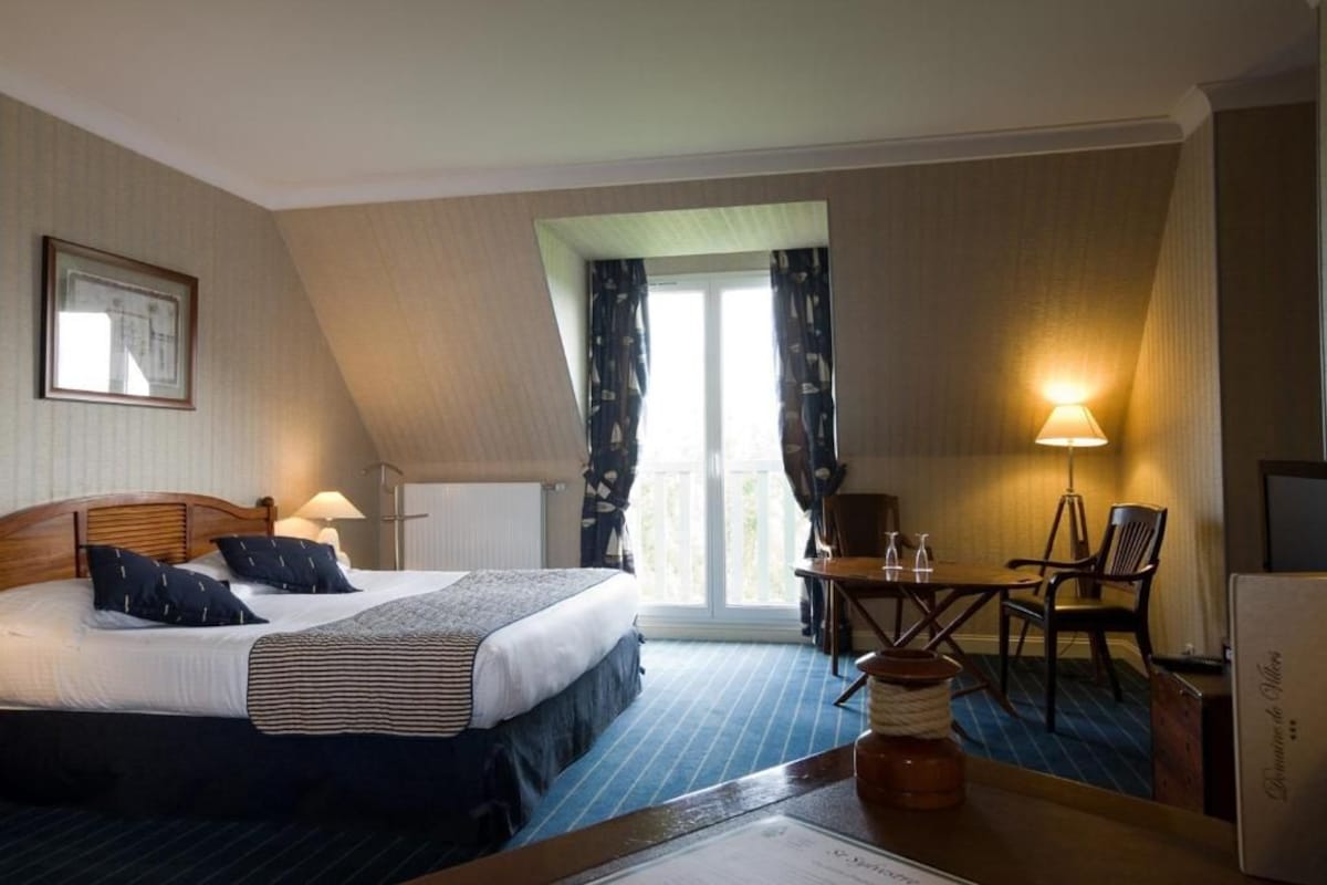 Ein Hotelzimmer mit Bett, Schreibtisch, Stuhl, Lampe und einem fensterbepflanzten Fenster.