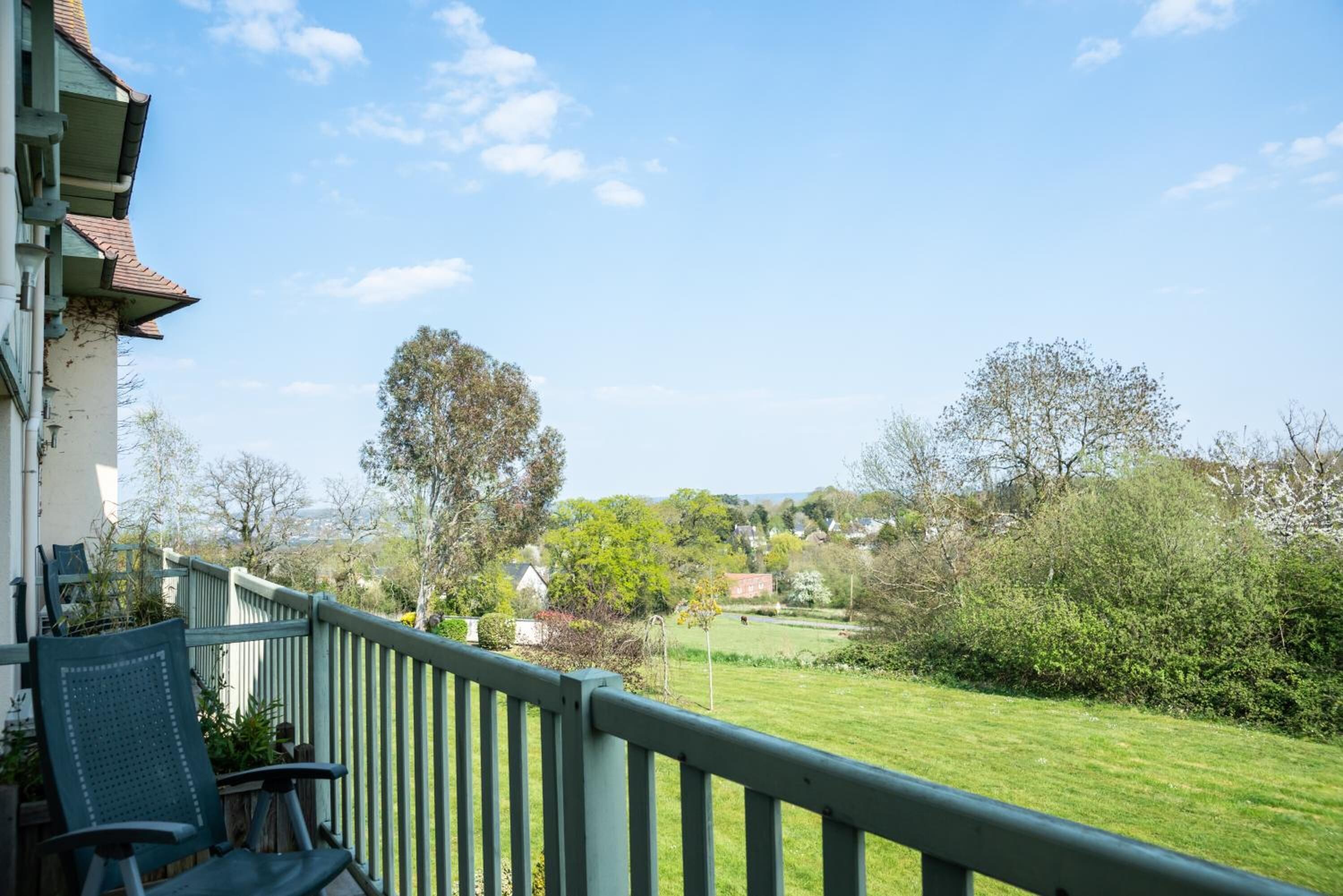 Un balcon doté d’une rambarde verte donnant sur un espace verdoyant, avec des arbres et des maisons visibles au loin.
