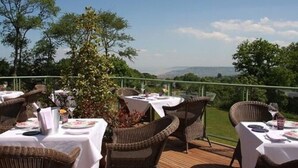 An outdoor dining area with wicker chairs and tables set up on a wooden deck, overlooking a scenic landscape with trees and hills.