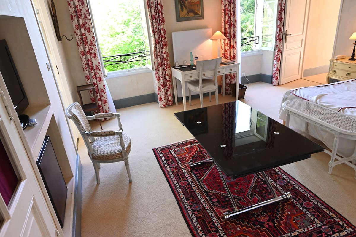 A bedroom with a bed, a desk, a chair, a window with curtains, and a view of greenery outside.