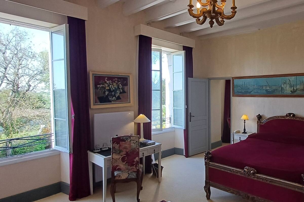 A bedroom with a large bed, a desk, a chair, a window with curtains, a chandelier, and a painting on the wall.