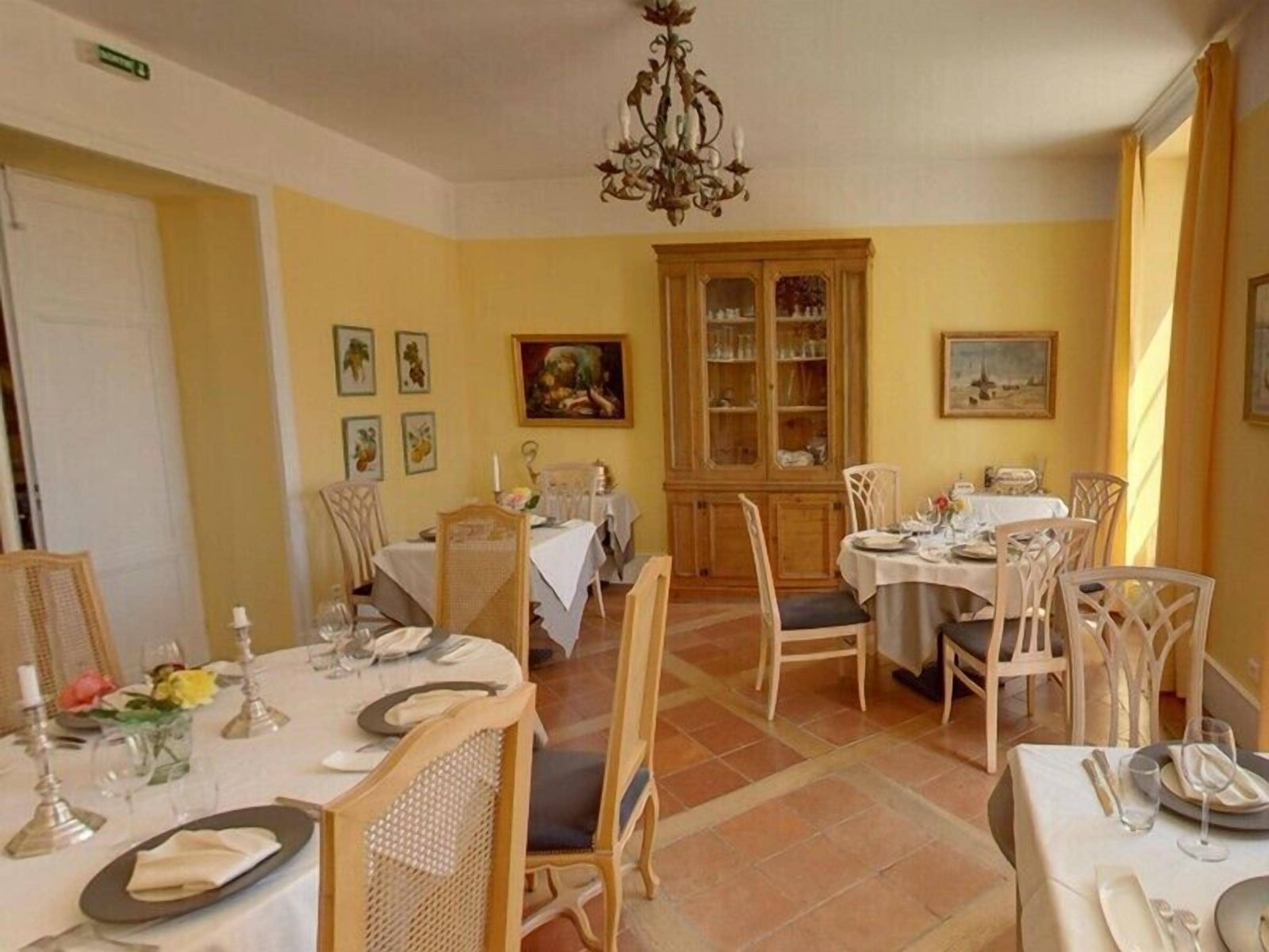 A dining area with multiple tables set for meals, wooden chairs, a chandelier, and a cabinet with glass doors.