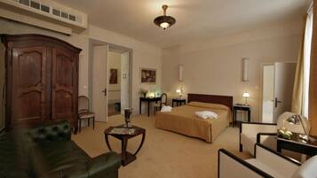 Una habitación de hotel con un gran armario de madera, un sofá de cuero, una cama con colcha color beige, dos mesitas de noche con lámparas y una pequeña mesa con un jarrón.