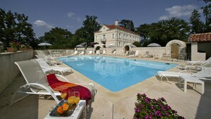 Zona de piscina con sillas reclinables y sombrillas, un edificio al fondo y cielo despejado.