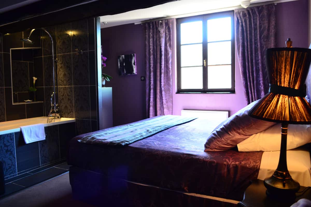 Une chambre à coucher comprenant un lit, une table de chevet, une lampe, une fenêtre avec des rideaux, et une salle de bain avec une douche et une baignoire.