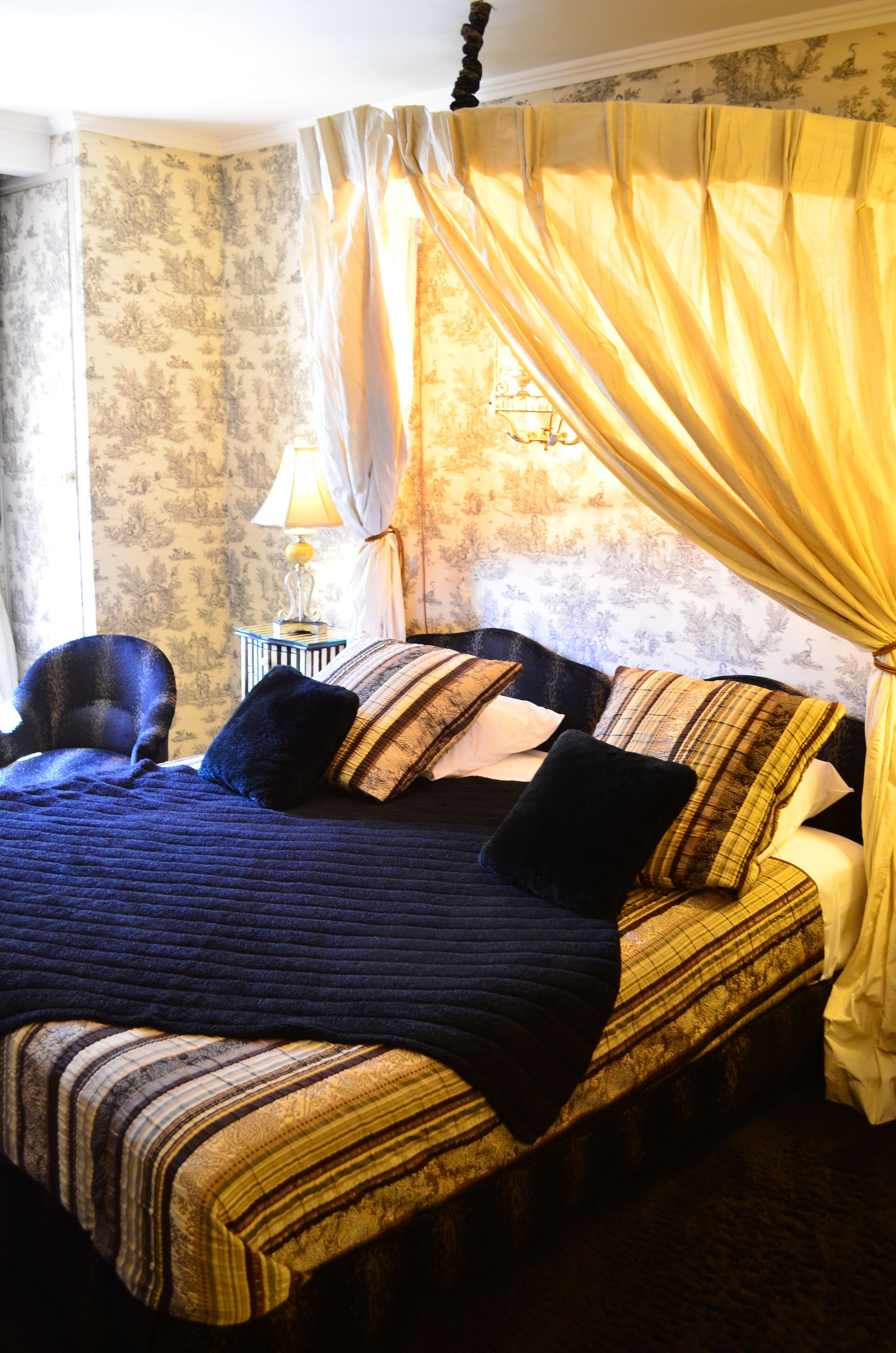Een bed met baldakijn, een lamp op een nachtkastje en een stoel.