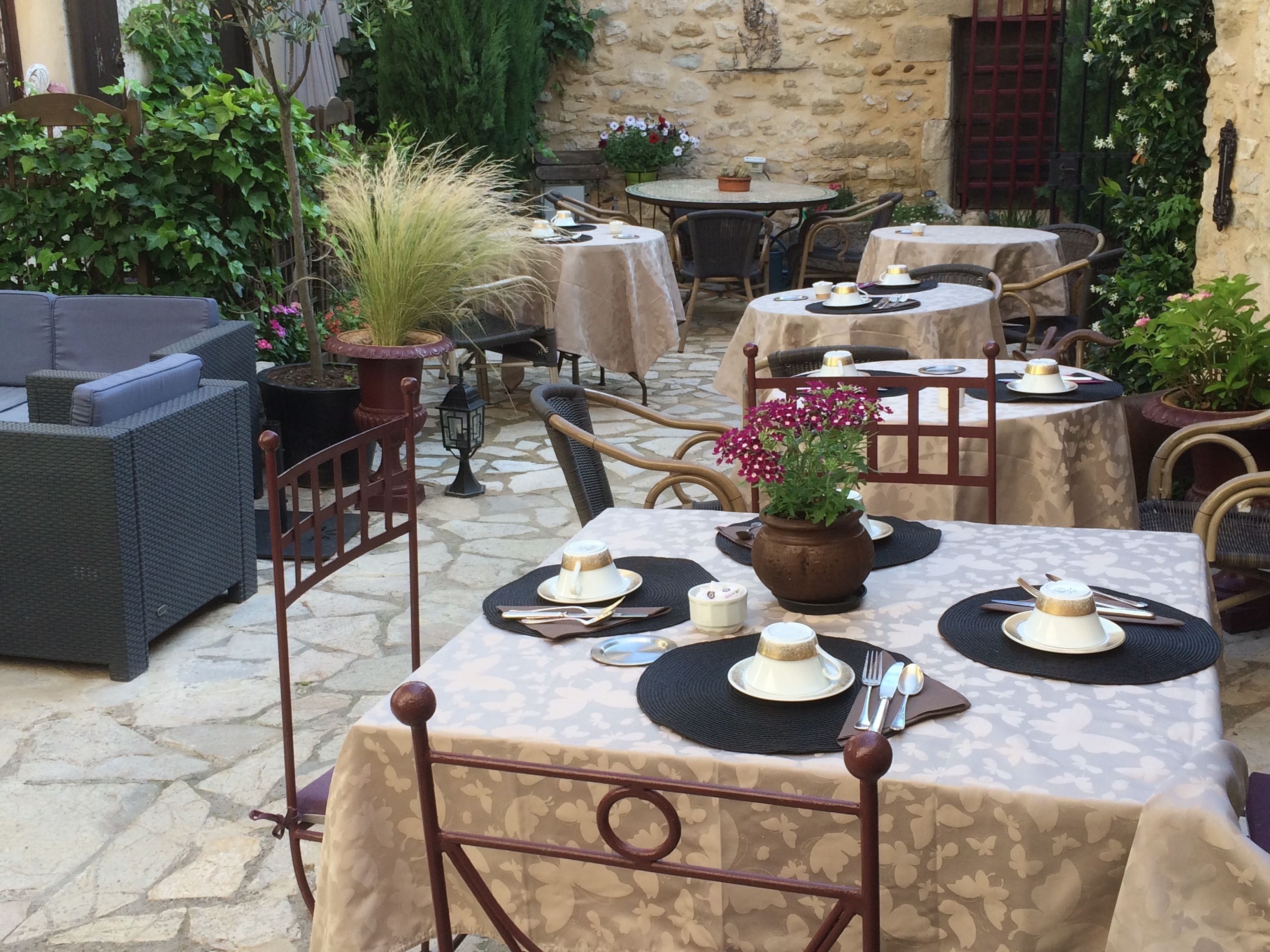 Une terrasse extérieure avec des tables et des chaises rondes, recouvertes de nappes, entourée de plantes en pot et d’un mur de pierre.