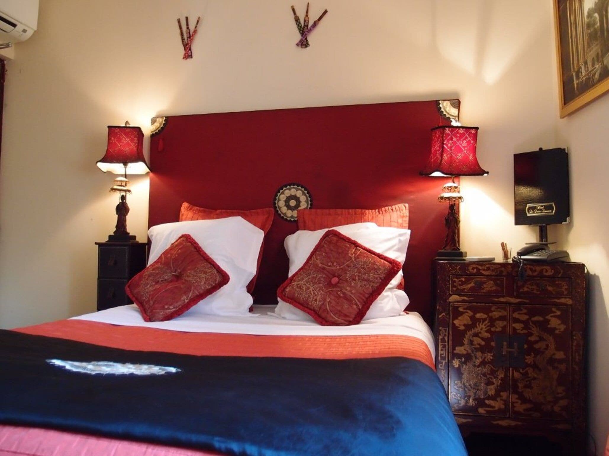 A bed with red and white bedding, two red lamps, a decorative wall hanging, and a wooden nightstand with a telephone.
