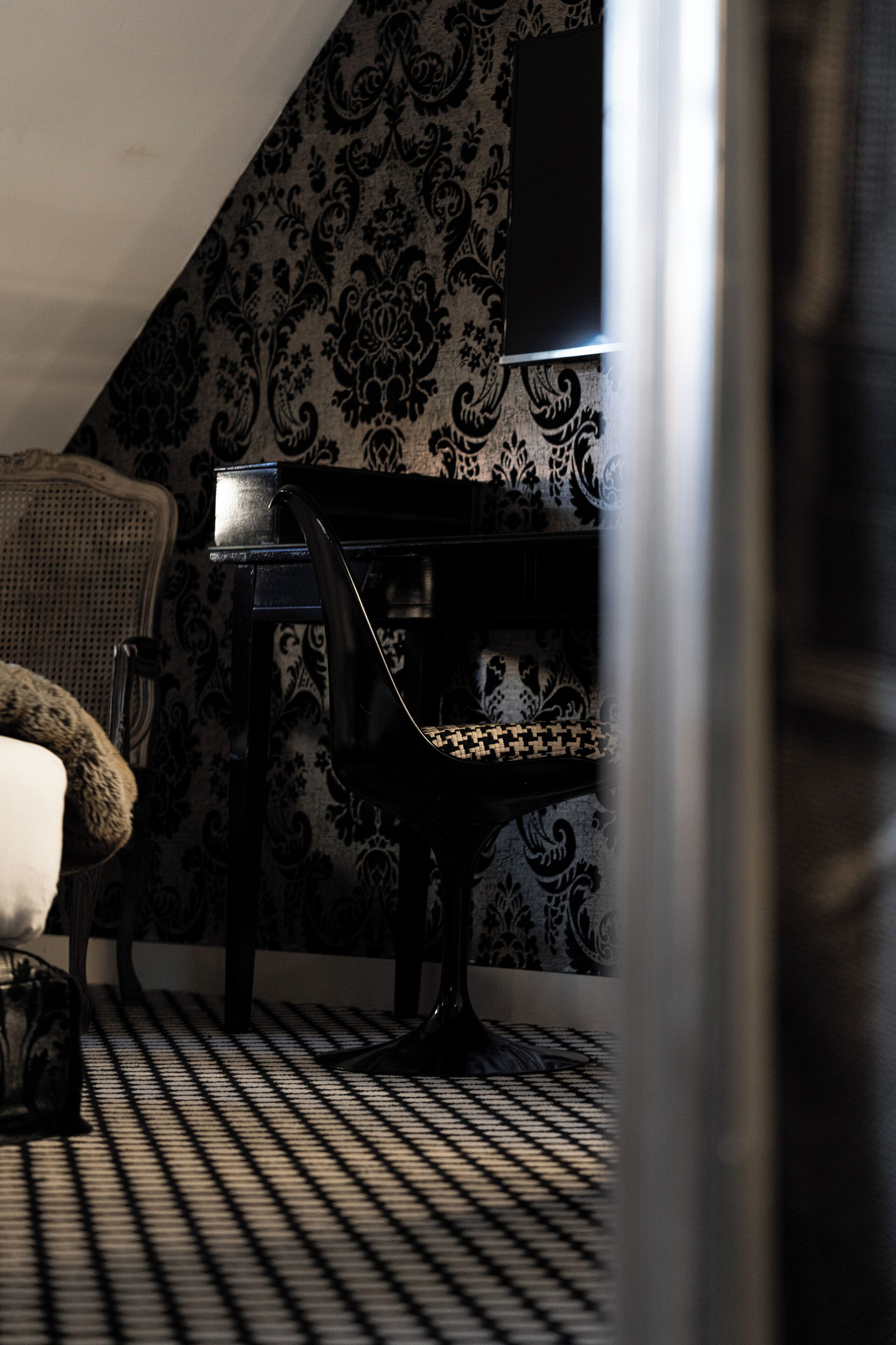 Una habitación con papel tapiz estampado, una silla negra con asiento estampado, una mesita y una lámpara.