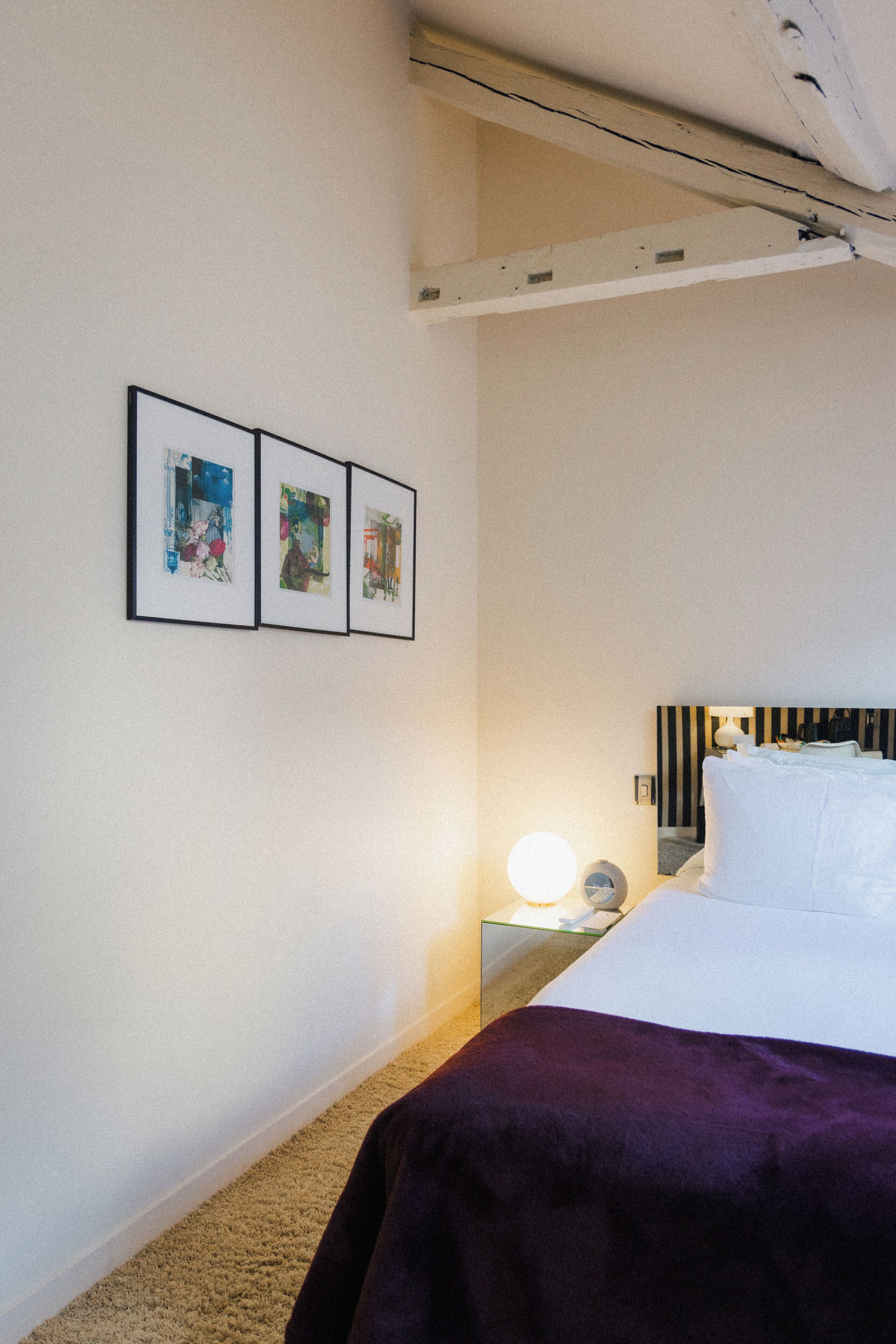 A hotel room with a bed, a nightstand with a lamp, and three framed artworks on the wall.