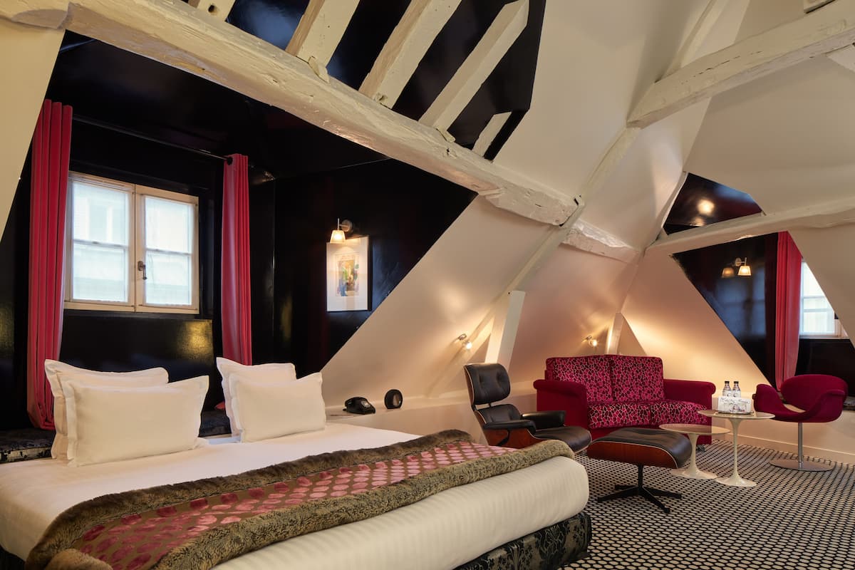 A bedroom with a bed, pillows, a red sofa, a red armchair, a small table with bottles, and a patterned rug.