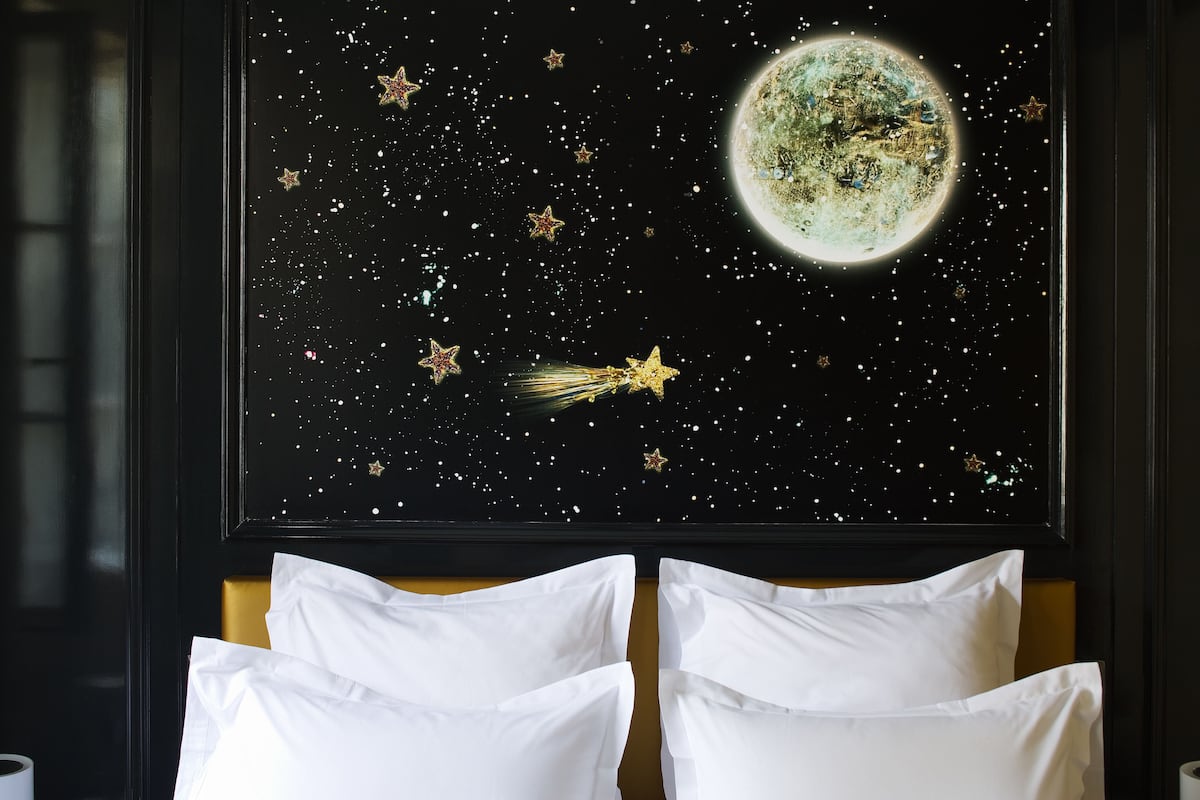 A bedroom with a bed against a wall, a framed picture of a starry night sky with a moon, and white pillows.