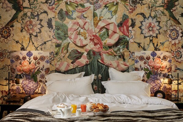 A bed with white linens, a floral headboard, and a breakfast tray with croissants, juice, and a coffee pot.