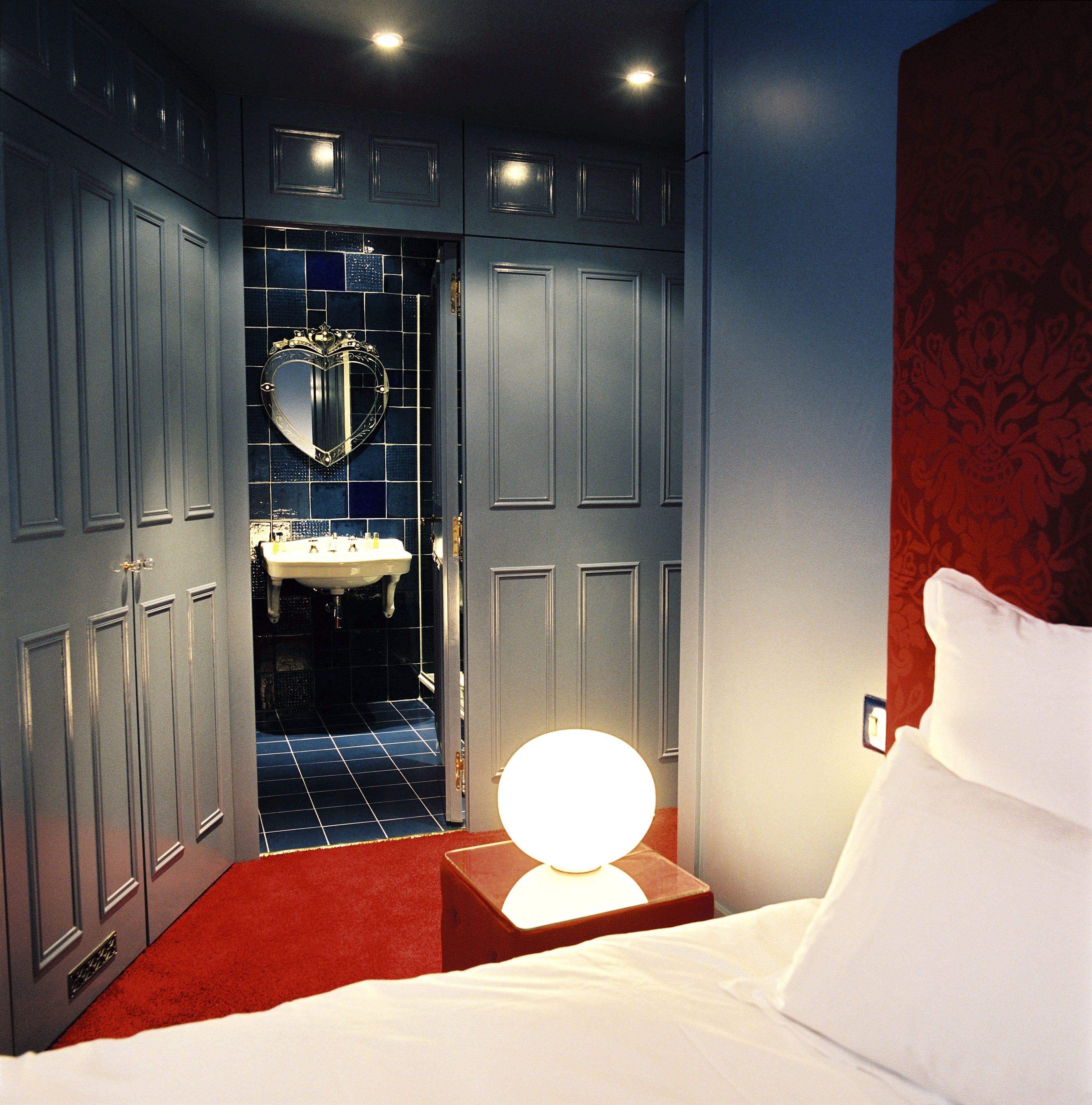 A hotel room with a bed, a nightstand with a lamp, a mirror, and a bathroom with a sink and tiles.