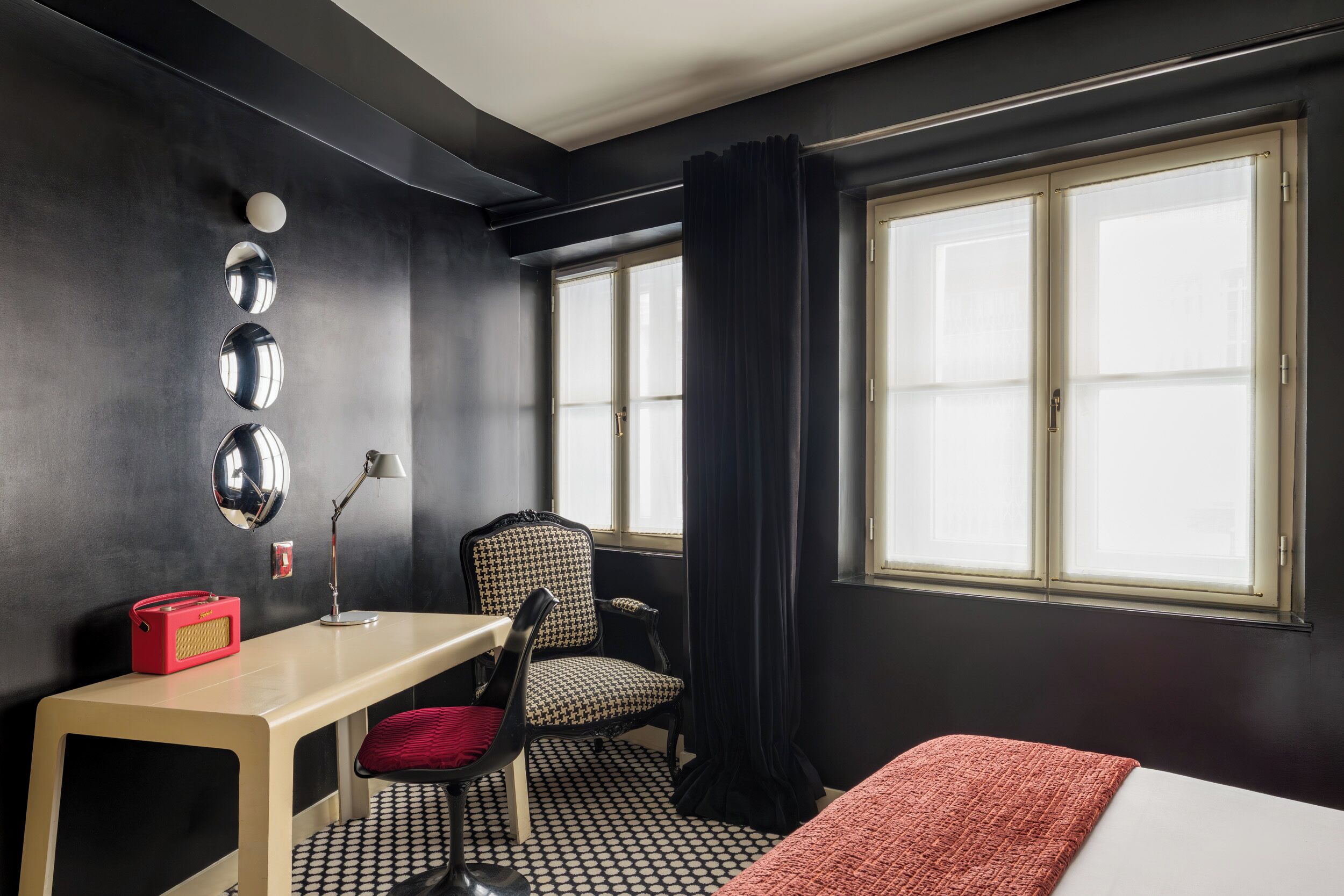 Une chambre d’hôtel avec un bureau, une chaise et un lit. De grandes fenêtres sont présentes, avec des rideaux, ainsi qu’un grand miroir rond accroché au mur et un sac rouge posé sur le bureau.