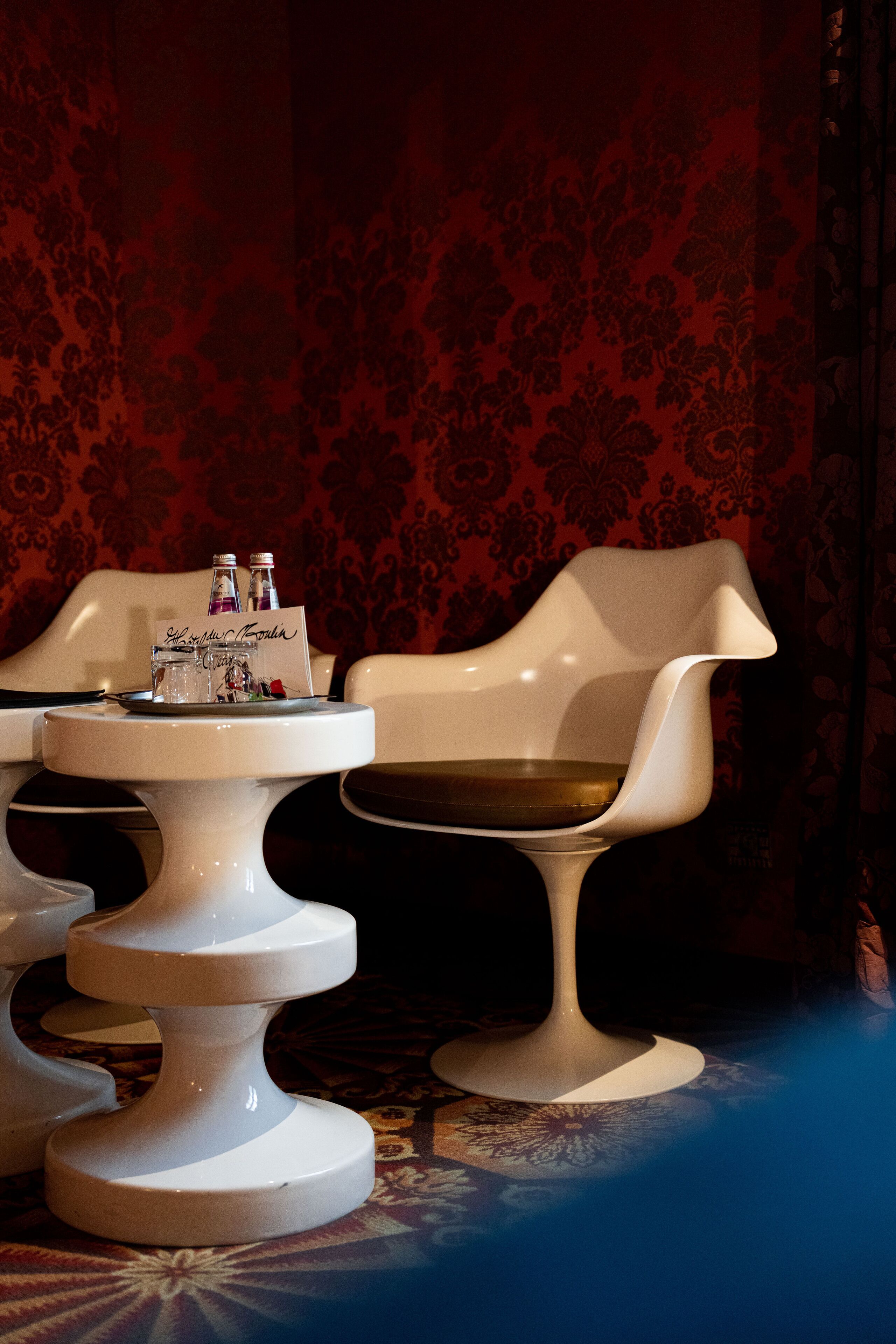 Une table blanche avec un plateau rempli de bouteilles et un panneau, une chaise blanche avec un coussin et un tapis à motifs.