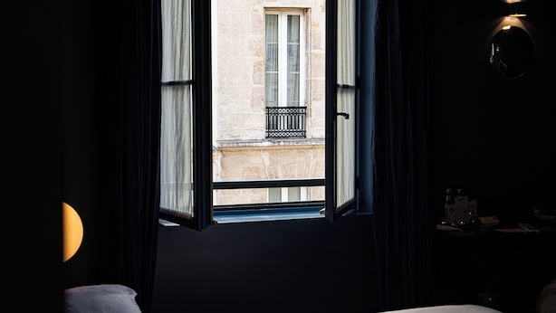 A hotel room with a bed, a window with a view of a building, and a nightstand with a lamp. - Hôtel du Petit Moulin, Haute Couture hotel by Lacroix (Paris)