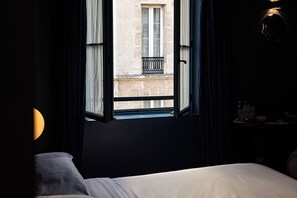 Habitación de hotel con cama, ventana que ofrece vistas a un edificio y mesita de noche con lámpara.