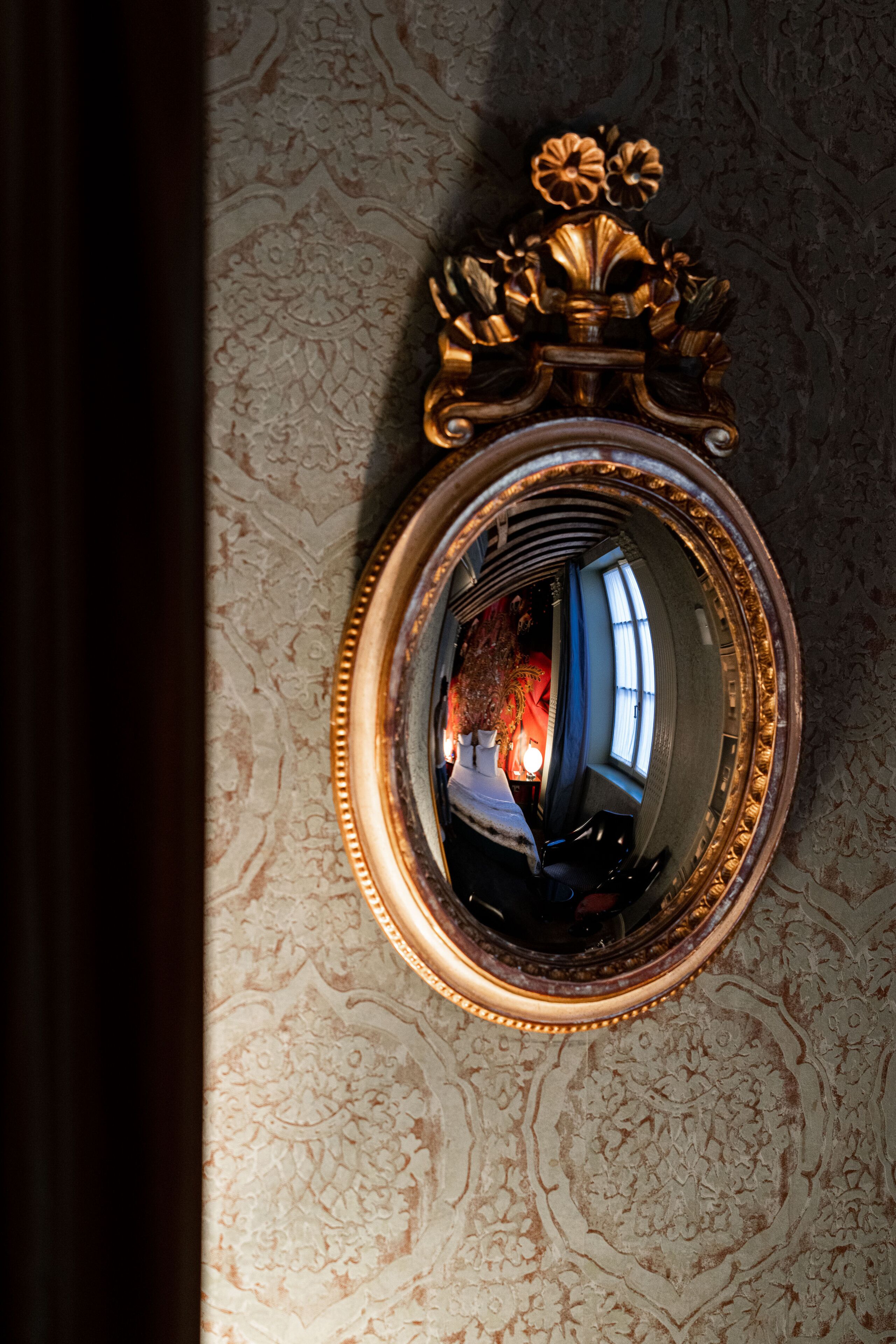 A mirror with an ornate gold frame reflecting a room with patterned wallpaper and a window.