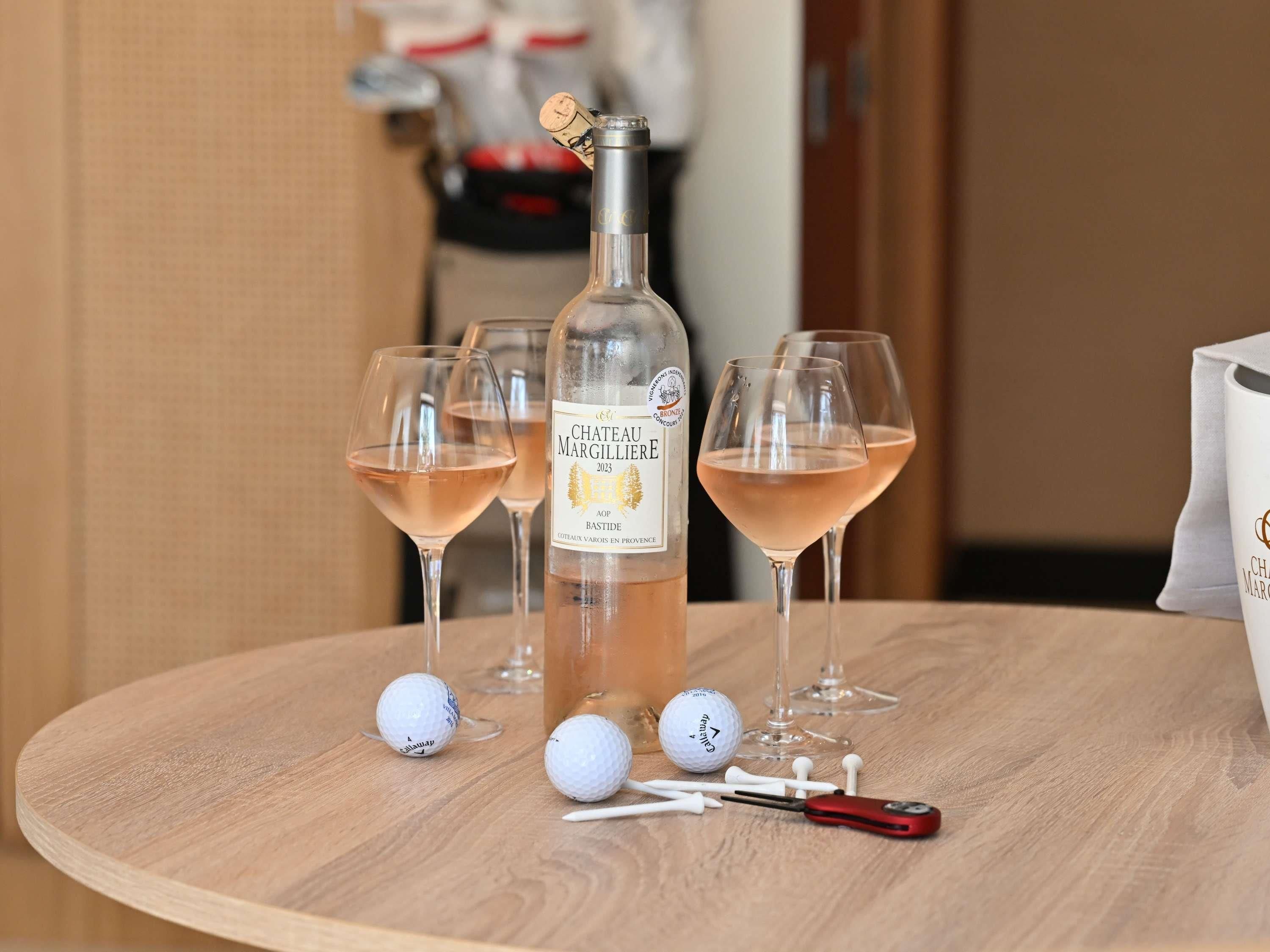 Eine Flasche Chateau Margillere 2023 AOP Bastide Wein, zwei Weingläser, Golfbälle und ein Golfschläger auf einem Holztisch.