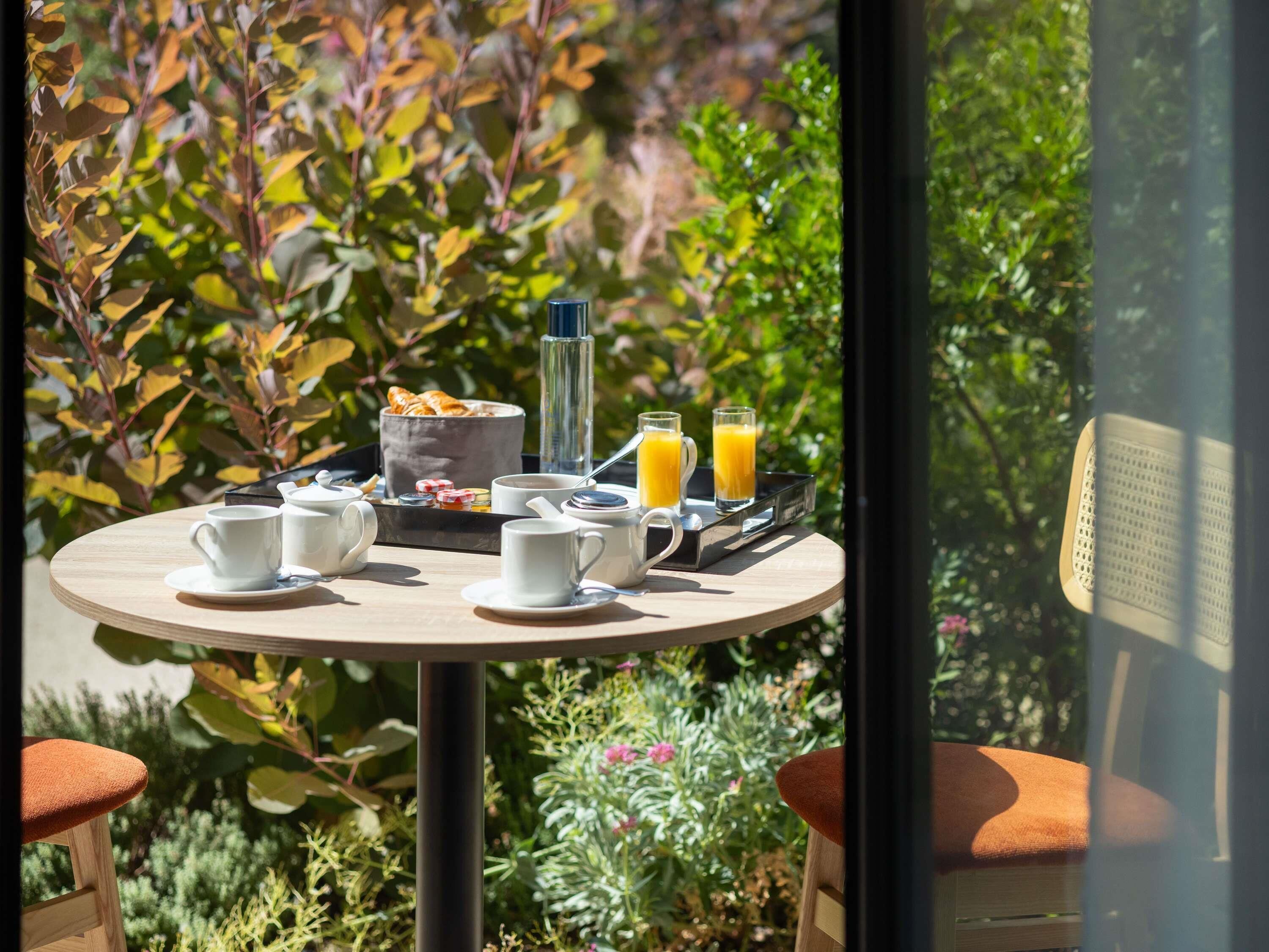 A breakfast setup with a tray of croissants, a coffee pot, cups, and orange juice on a table outdoors.