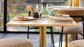 A dining table with plates, glasses, and a bowl of bread, set against a window with a view of greenery.
