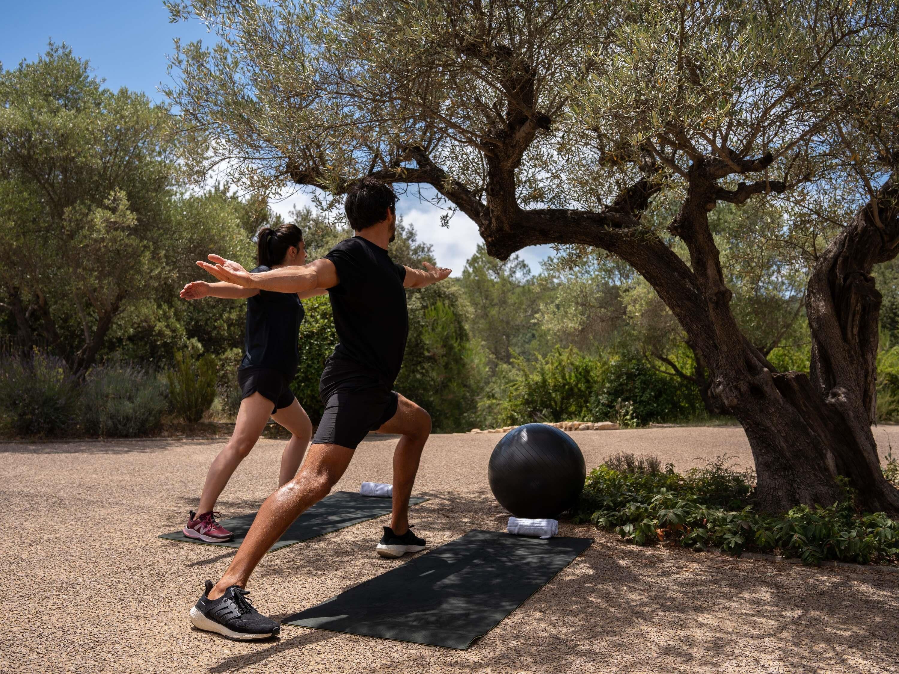 Deux personnes font de l’exercice en plein air sur un chemin de gravier, bordé d’arbres, et utilisent un grand ballon d’exercice.