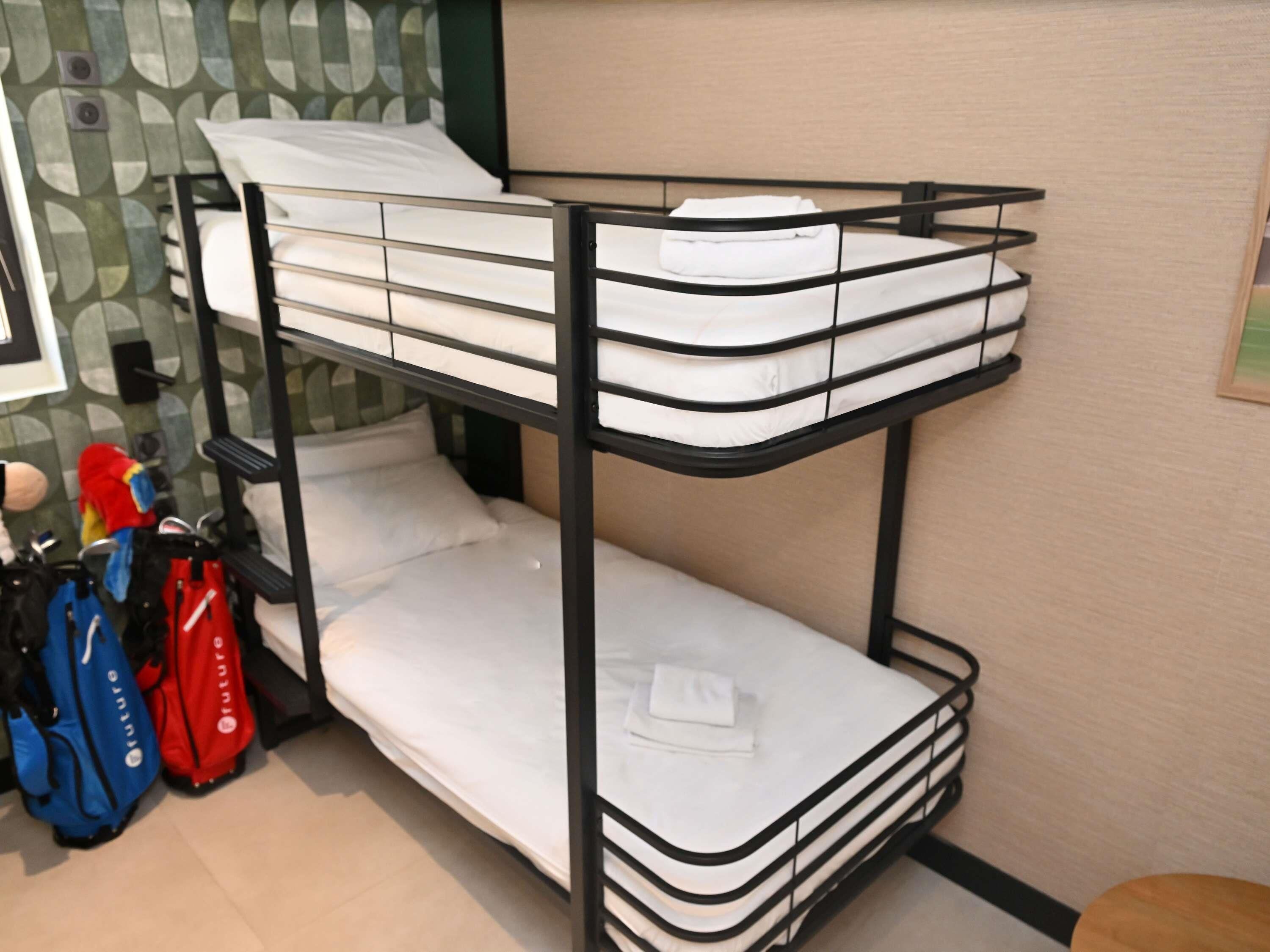 A bunk bed with white bedding and a black metal frame. There are two bags on the floor beside the bed.