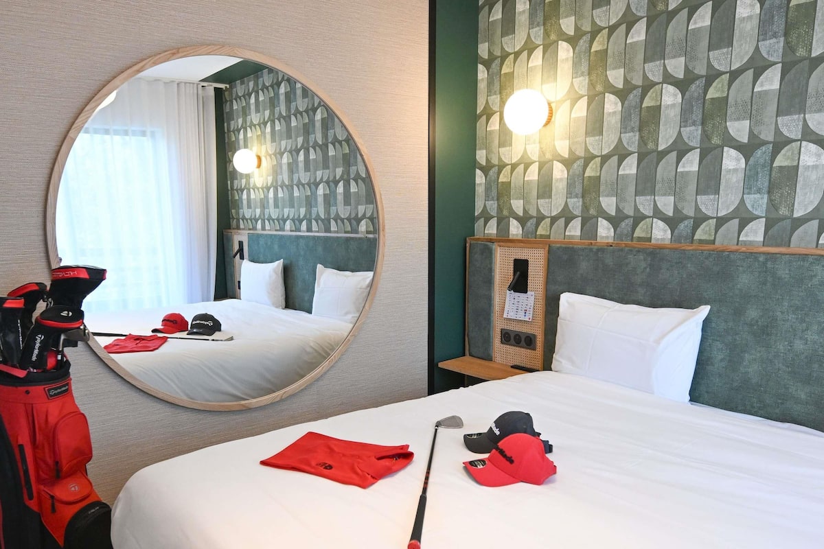 A hotel room with a round mirror, a bed with white linens, a golf bag, and a red cap on the bed.