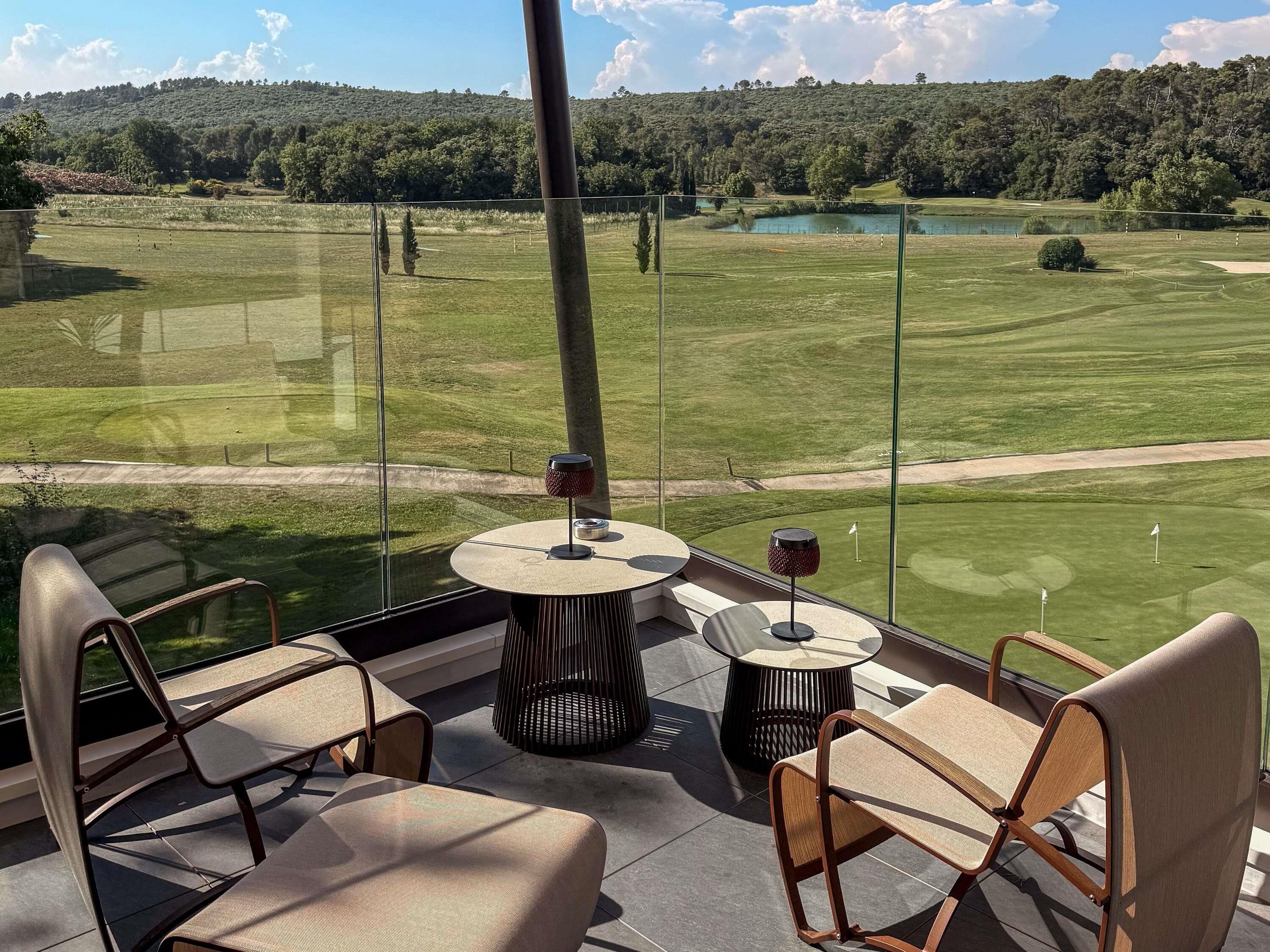 A balcony with a view of a golf course, a round table with chairs, and a glass railing.