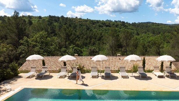 A poolside area with sun loungers and umbrellas, surrounded by a forested hillside.