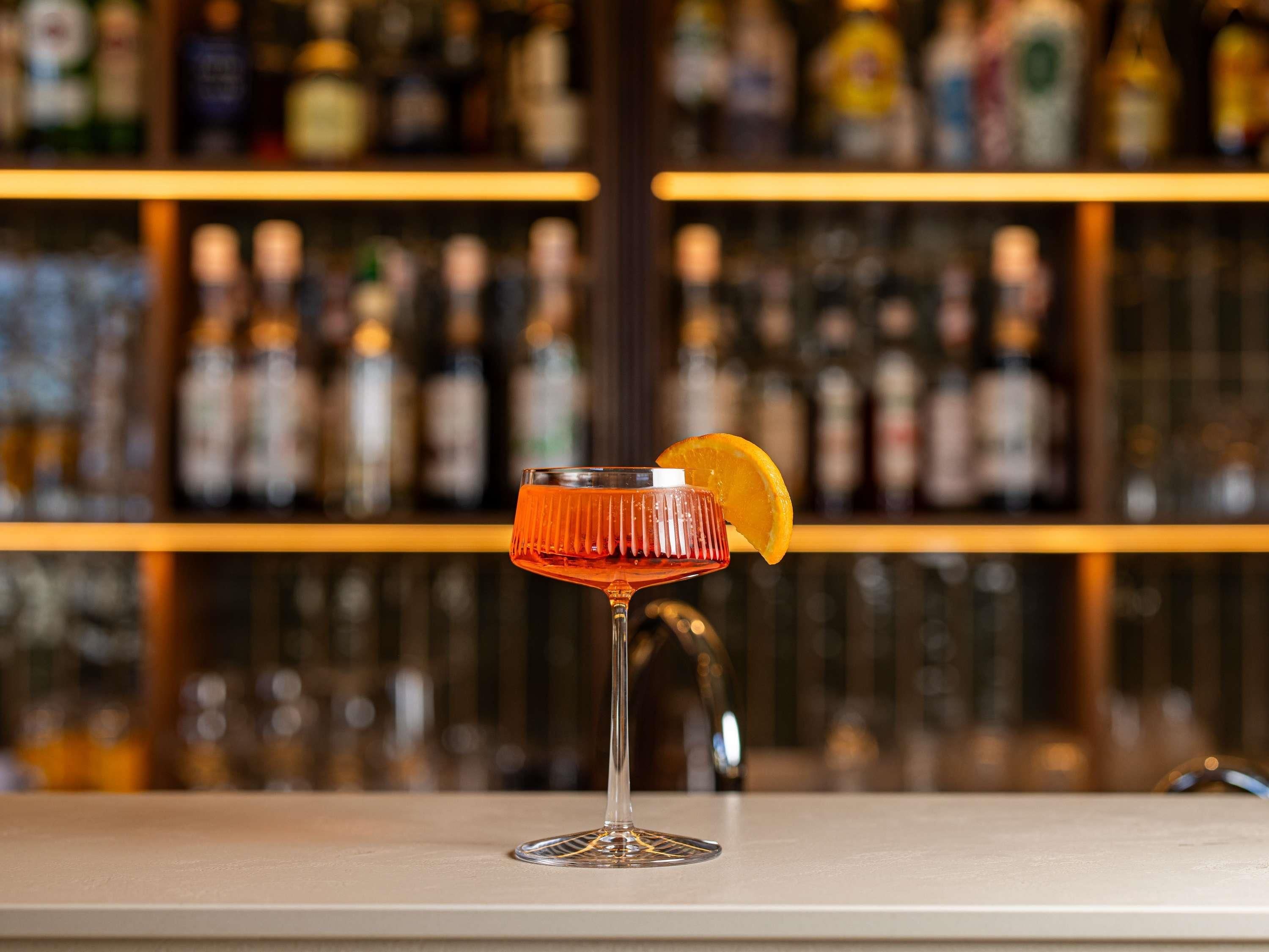 A bar setting with a cocktail glass on a counter, a bottle of liquor behind it, and shelves with various bottles in the background.