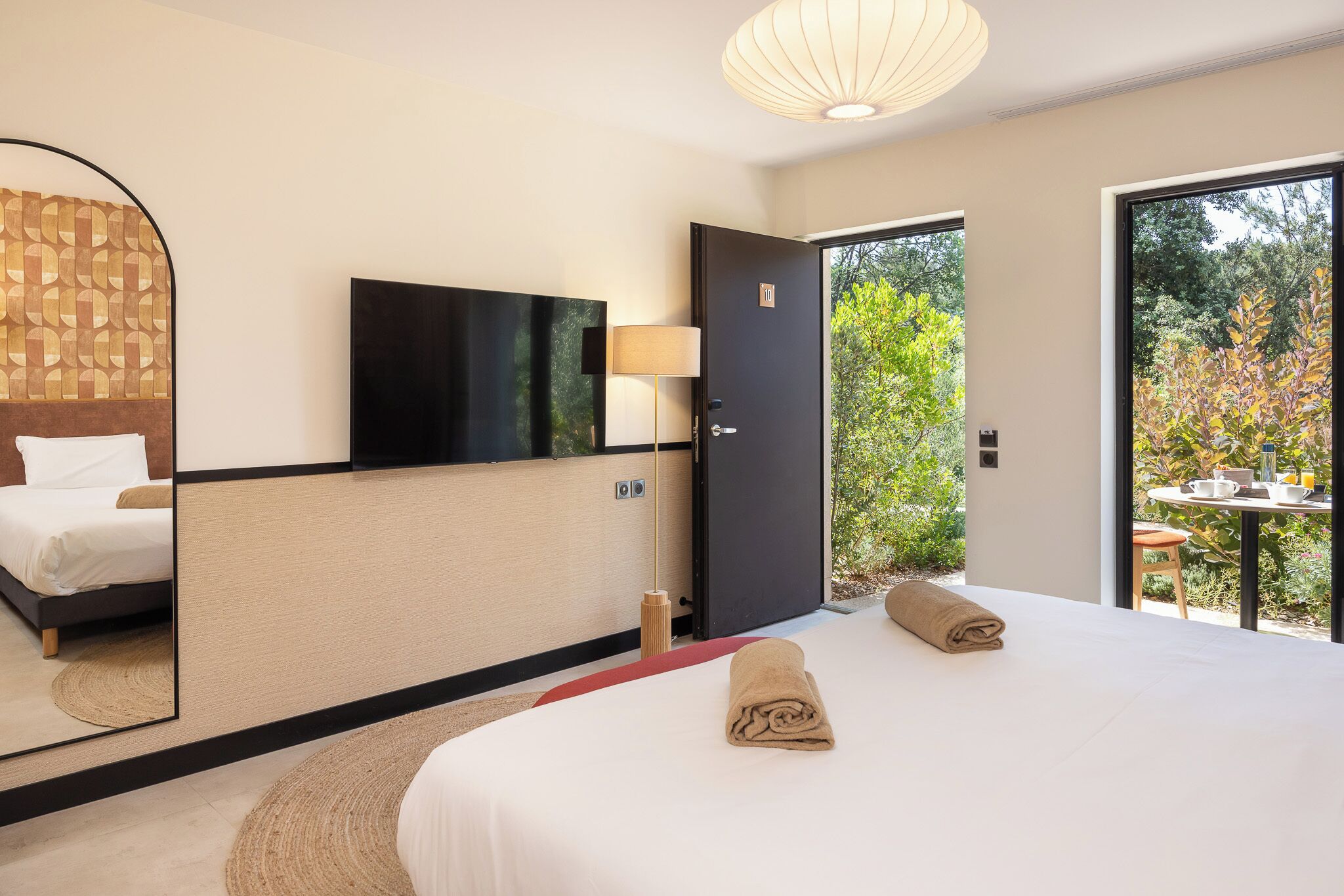 Habitación de hotel moderna con una cama grande, televisión de pantalla plana, un espejo, una puerta que da a un espacio exterior y una pequeña mesa con una silla.