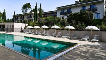 Una piscina con sillones de descanso y sombrillas, rodeada de edificios y vegetación.